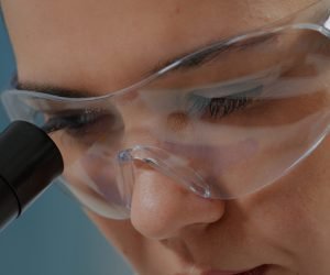 Woman chemist with protective glasses using microscope in laboratory. Microbiologist working with microscopic lens on optical tool to analyze genetic substance with magnifying glass. Close up